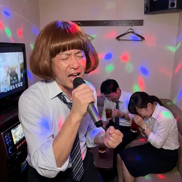 A man in a shirt and tie belting karaoke in the bob while his colleagues cry laughing